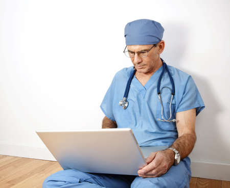 Mature doctor sitting on floor, leaning against wall, in blue scrubs, working on his laptopの写真素材
