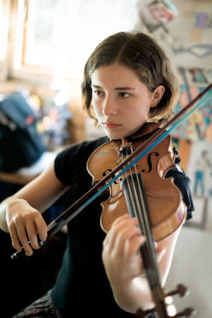 Pretty young woman practicing violin in a sunny room. Available light, selective focus, focus sharpest on eyes.の写真素材