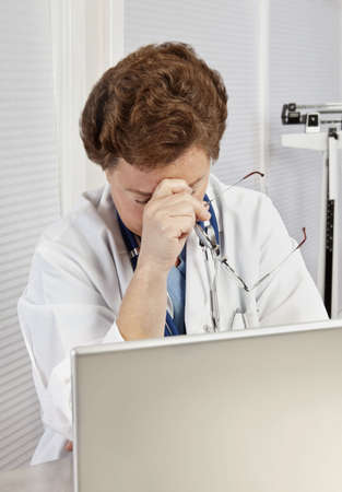 Mature female doctor or nurse in blue scrubs and lab coat, working on laptop, rubbing her tired eyes, looking overworked and worried.の写真素材