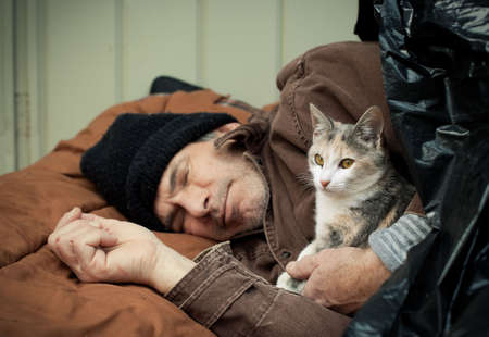Closeup portrait of a homeless older man sleeping under a plastic tarp on the street with a friendly stray kitten. Selective focus on the man's hands and the kitten.の写真素材