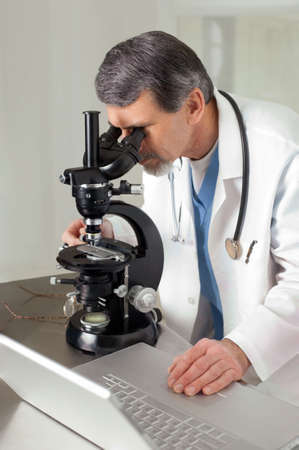 A male doctor or scientist looking through a microscope on a table with laptop computer.の写真素材