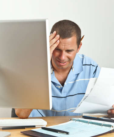 Worried young man holds his head in pain as he wonders how he will pay the bills. Seated at desk with computer, checkbook, stacks of bills and calculator.の写真素材