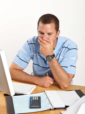 Worried young man, stressed out over bills and finances, sitting at desk with checkbook and computer.の写真素材