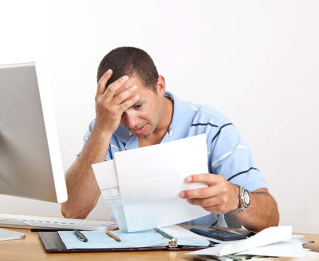 Young man at desk with computer and checkbook, worrying about paying bills, holding his head.の写真素材