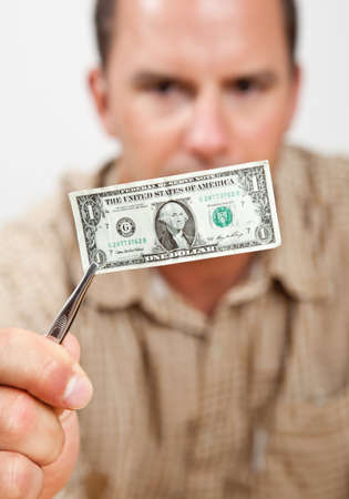 Man holding tiny shrinking dollar with tweezers. Short depth of field. Man's hand, tweezers and dollar bill are in sharp focus, man's face and shirt are blurry...intentionally.の写真素材
