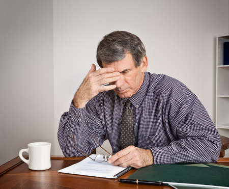 Tired, stressed out businessman rubs his eyes while seated at a desk reading reports.の写真素材