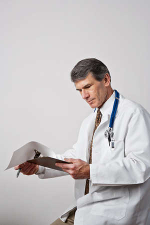 Handsome mature male doctor reviewing his notes with clipboard and pen. Plain gray background. の写真素材