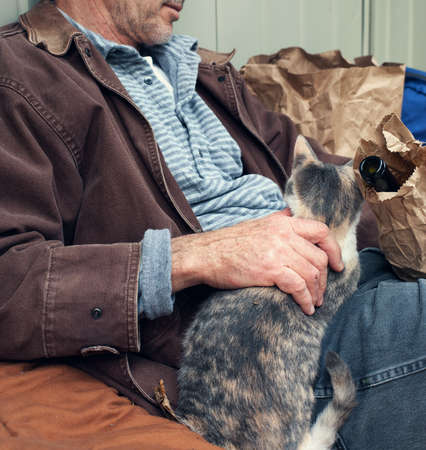 Homeless man outdoors, seated, with wine bottle in paper sack and friendly stray cat  Square format  Sharpest focus on manの写真素材