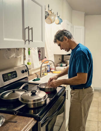 Candid lifestyle shot of a mature man cooking dinner alone in a plain small kitchen. Man is backlit, high speed shot with wide aperture, some grain evident.の写真素材