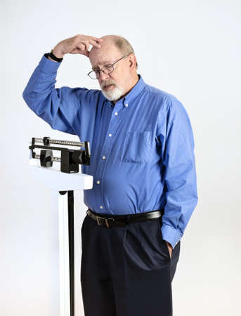 Senior caucasian man weighing himself on vertical weight scale, while scratching his head in confusionの写真素材