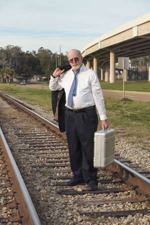 A senior businessman in suit and tie carrying his old suitcase, walks down a railroad track, tired, defeated, and jobless with no prospects. Nearby interstate overpass adds further symbolism as he is on an outmoded railroad track, life having passed him bの写真素材