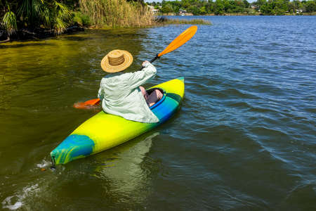 Woman in a straw hat paddles away from shore in her kayak on a beautiful river or lake.の写真素材