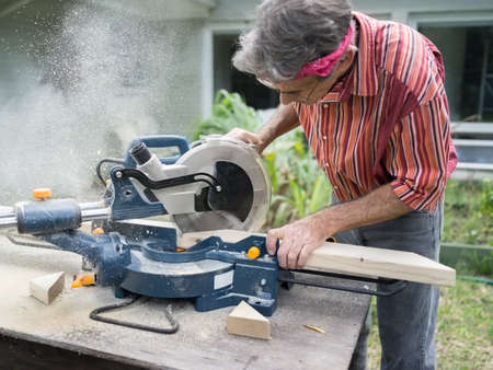 Closeup of mature man sawing lumber with sliding compound miter saw outdoors, sawdust flying aroundの写真素材