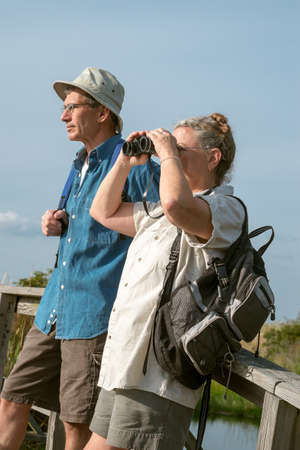 Happy mature man and woman resting on an old wooden footbridge, watching birds with binoculars. Short depth of field, woman in sharpest focus.の写真素材