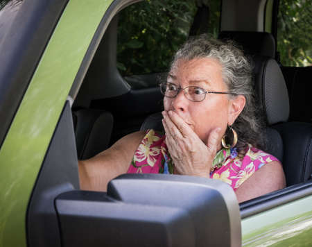 Sweaty, hot, senior woman driver looking terrified holding hand to mouth in driver