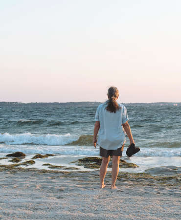 Back view of barefoot, middle aged woman on a seaweed laden beach looking out over the surf as the day dawns.の写真素材