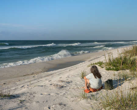 Backview of woman seated on sand dunes at Florida beach, gazing out to sea at waves and crashing surf.の写真素材