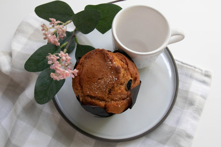 A warm, inviting scene featuring a golden-brown muffin wrapped in black paper, set beside a white teacupの写真素材