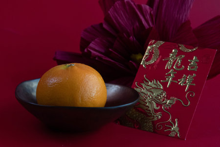A festive still life featuring a ripe orange resting in a dark bowl beside a traditional red envelope (hongbao) embossed with golden dragon motifs and auspicious Chinese charactersの写真素材