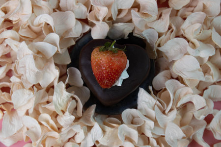 A rich, heart-shaped chocolate dessert topped with a fresh red strawberry and a touch of whipped cream, nestled among soft dried petals.の写真素材