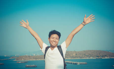 handsome asian young man smiling and  raising his hands , with blue sea and sky backgroundの写真素材