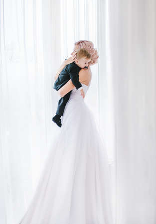 Beautiful bride in white dress and her son in white roomの写真素材