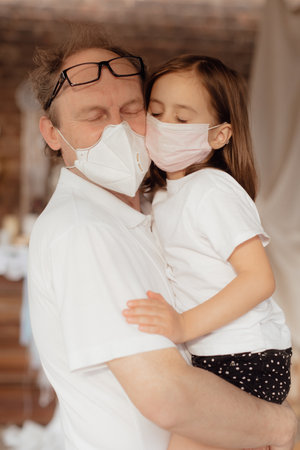 Stock Photo - Little granddaughter in a face mask hugs and wants to protect grandfather from an epidemic. Family support during quarantine isolation due to outbreaks of coronavirus. Elderly at riskの写真素材
