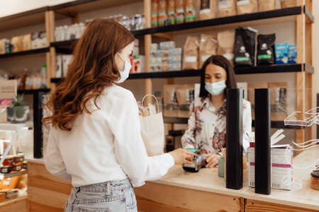 Girl buyer in a mask makes a purchase in the store. Saleswoman or small business owner wearing medical mask at the counter in cafe or small shop. Concept of a retail business during a pandemic.の写真素材