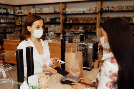 Girl buyer in a mask makes a purchase in the store. Saleswoman or small business owner wearing...