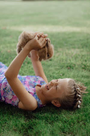 Little girl sitting on the green grass with rabbit. Cute child girl holding a bunny in her hands on Easter day. Fluffy brown plush pet.の写真素材