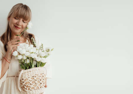 Ederly woman with flowers, she wearing light clothes on a white background. Portrait senior woman with place for text. Elderly lifestyle at home concept. Mock-up for design.の写真素材