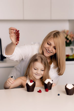 Pastry chef confectioner young caucasian woman with her daughter decorate a cake and cupcakes in the kitchen.の写真素材