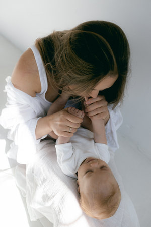Young woman with baby in her arms. Blonde girl in white clothes smiling to your child on white background. Happy motherhood and breastfeeding concept. Photos with sun glare, soft focus, overexposure.の写真素材