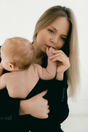 Young woman with baby in her arms. Blonde girl in black clothes smiling to your child on white background. Happy motherhood and breastfeeding concept. Photos with sun glare, soft focus, overexposure.の写真素材