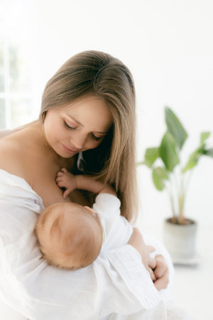 Young woman with baby in her arms. Blonde girl in white clothes smiling to your child on white background. Happy motherhood and breastfeeding concept. Photos with sun glare, soft focus, overexposure.の写真素材