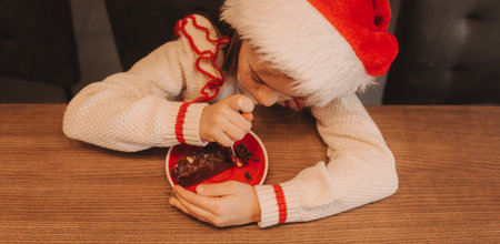 Cute funny schoolgirl wearing a pullover and Santa Claus hat, smiling, eating a chocolate dessert at home. Festive New Year food concept.の写真素材