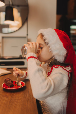 Cute funny schoolgirl wearing a pullover and Santa Claus hat, smiling, eating a chocolate dessert at home. Festive New Year food concept.の写真素材
