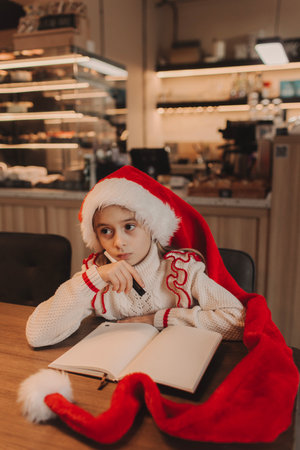 Smiling joyful girl in Santa hat writes wishes in notepad while sitting in cafe during winter holidays on New Year's Eve. Soft focusの写真素材