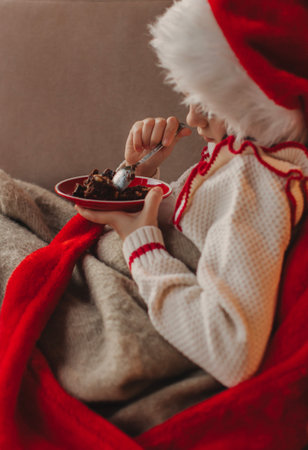 Cute funny schoolgirl wearing a pullover and Santa Claus hat, smiling, eating a chocolate dessert at home. Festive New Year food concept.の写真素材