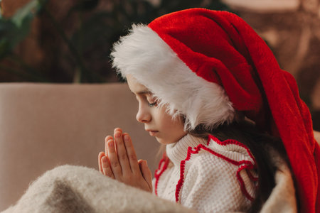 Close-up Caucasian child, lovely baby girl in Santa hat, makes cherished wish and cute presents, surrounded by Christmas decorations. Magic festive atmosphere at home.の写真素材