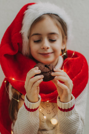 A cute teenage girl in a pullover and Santa Claus hat holds and eats a chocolate cookie against the background of a light wall. Festive New Year food concept.の写真素材
