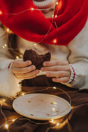 Hands of a teenage girl holding a cookie against the background of a Santa hat and garland lights. Festive New Year food concept. Close-up photo.の写真素材