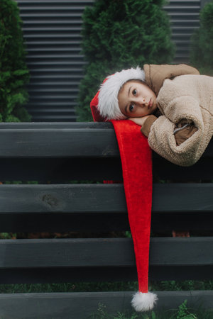 Adorable teen girl in Santa hat near Christmas tree waiting for Santa Claus outdoors. Girl against the background of a black fence with green fir trees. Celebration seasonal winter holidays with kids.の写真素材