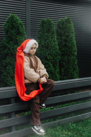 Adorable teen girl in Santa hat near Christmas tree waiting for Santa Claus outdoors. Girl against the background of a black fence with green fir trees. Celebration seasonal winter holidays with kids.の写真素材