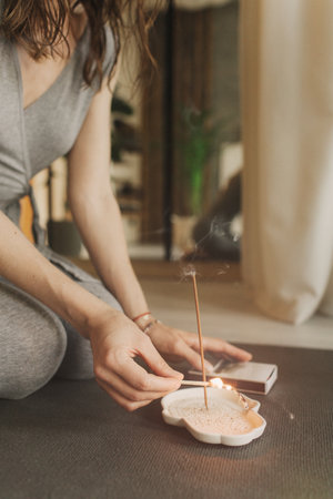A young woman lighting incense sticks an home before yoga. Buddhist healing practices. Clearing the space of negative energy. Aromatherapy. Selective focus.の写真素材