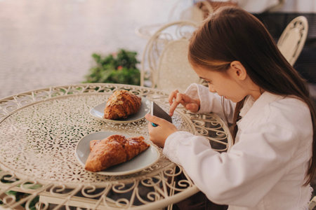 Cheerful preschool girl having breakfast and plays on the phone in the cafe on the city street. Concept of childhood and gadgets, networking.の写真素材