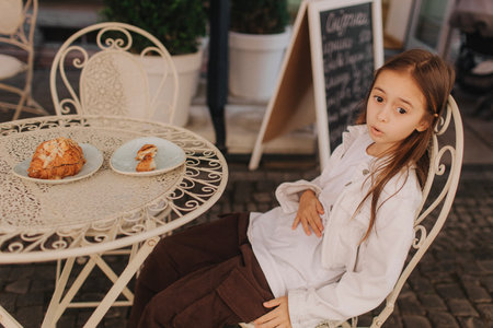 Dissatisfied capricious preschool girl having breakfast in the cafe on the city street. The teen is eating a croissant and looks into the camera. The concept of overeating and healthy eating.の写真素材