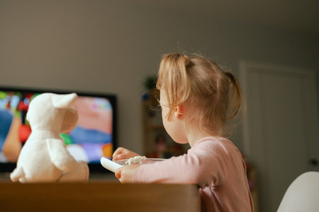 Baby girl eats breakfast and watches cartoons on TV. Children and technology concept. Alpha Generation and modern technology with electronic device. Part of the series.の写真素材