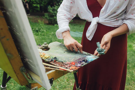 Close up of female artist's hands clear the palette of paints on the easel. Mindfulness, art therapy, creativity concept. Process of plein air painting in nature. Part of the series.の写真素材