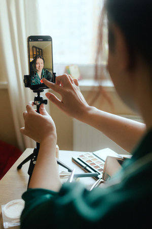 Plus size woman applying makeup on face with brush while recording vlog on smartphone for social...
